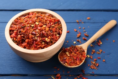 Photo of Chili pepper flakes in bowl and spoon on blue wooden table, closeup