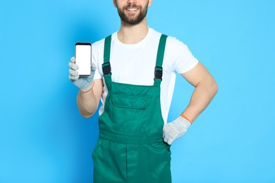 Professional repairman in uniform showing smartphone on light blue background, closeup