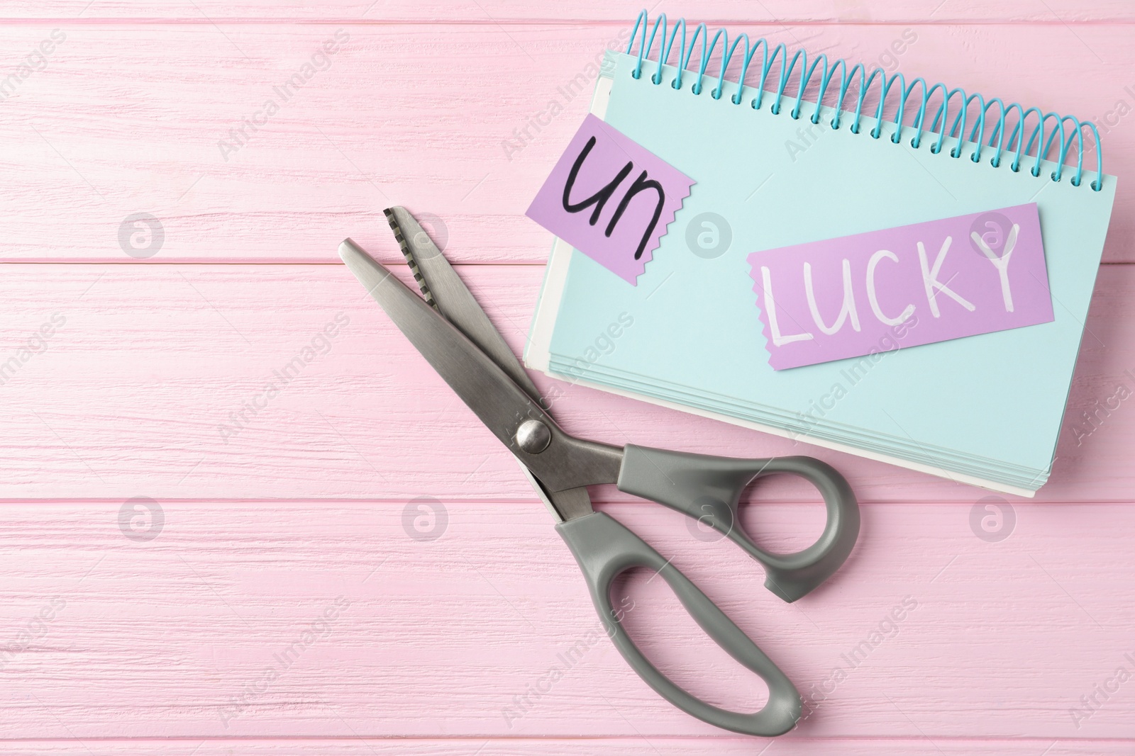 Photo of Sheets of paper with word UNLUCKY, notebook and scissors on pink wooden table, flat lay. Space for text