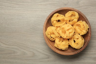 Raw tagliatelle pasta in bowl on light grey wooden table, top view. Space for text