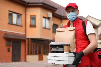Courier in protective mask and gloves with orders near house outdoors. Food delivery service during coronavirus quarantine