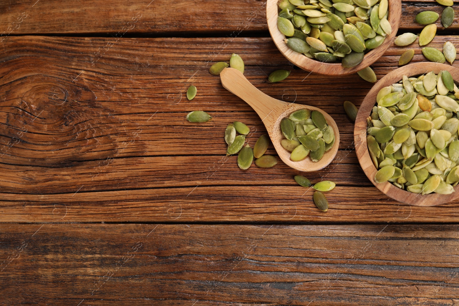 Photo of Bowls and spoon with peeled pumpkin seeds on wooden table, flat lay. Space for text