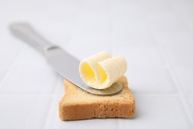 Photo of Tasty butter curl, knife and piece of dry bread on white tiled table, closeup