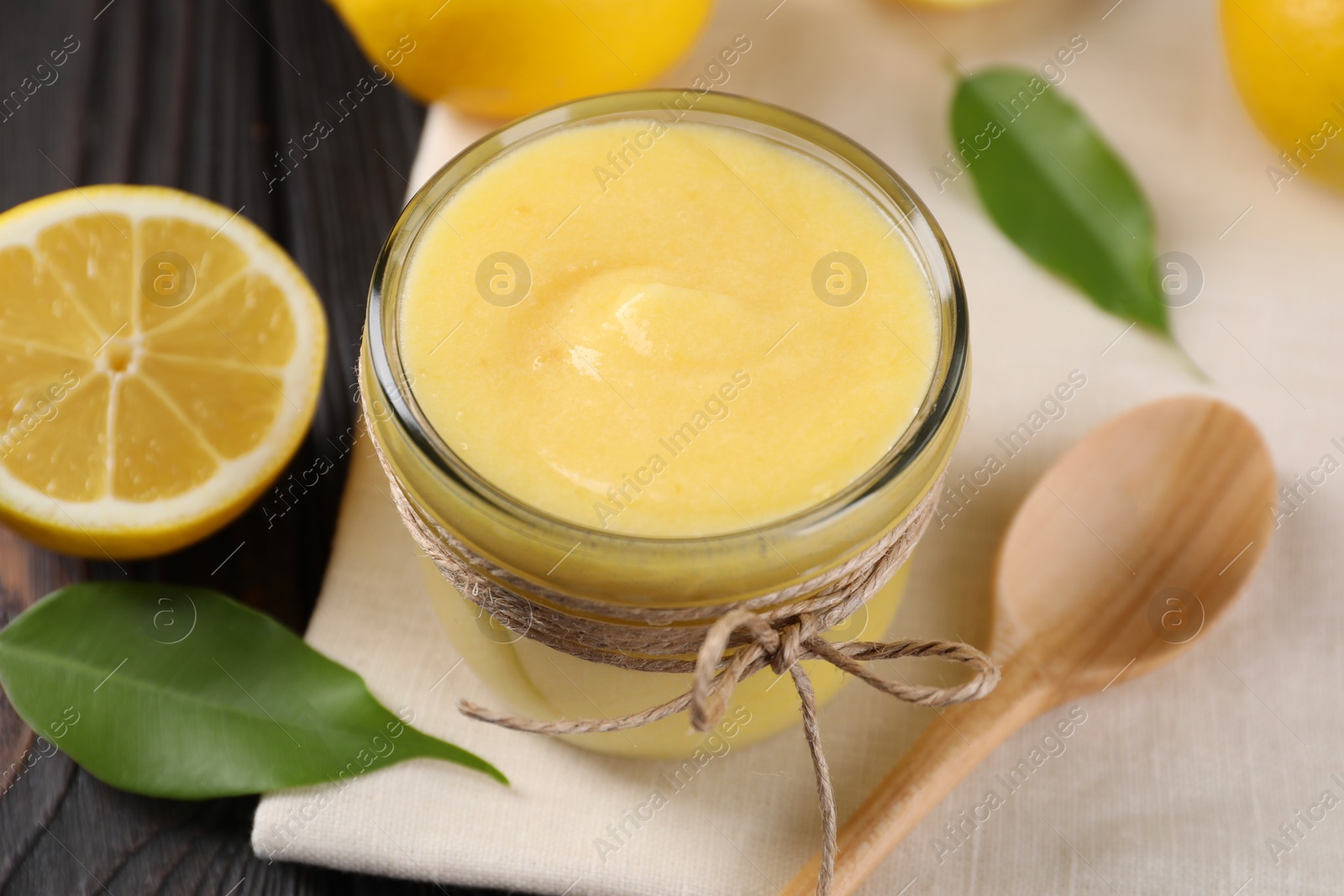 Photo of Delicious lemon curd in bowl, fresh citrus fruit, spoon and green leaves on wooden table, closeup