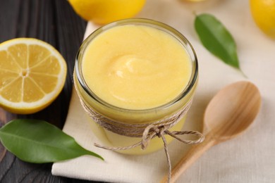 Delicious lemon curd in bowl, fresh citrus fruit, spoon and green leaves on wooden table, closeup