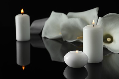 Photo of Burning candles and white calla lily flowers on black mirror surface in darkness, closeup with space for text. Funeral symbols