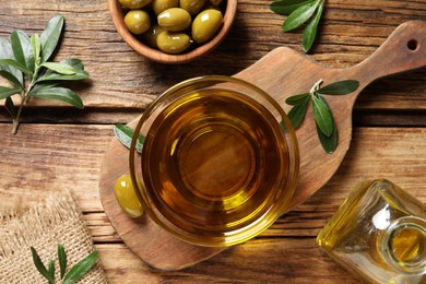 Photo of Glass bowl of fresh oil, ripe olives and green leaves on wooden table, flat lay