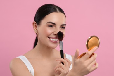 Beautiful woman with cosmetic pocket mirror applying makeup on pink background