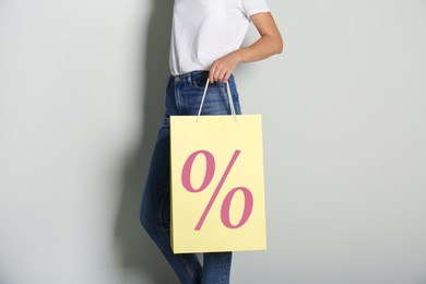 Image of Woman holding paper shopping bag with percent sign on light grey background, closeup. Discount concept