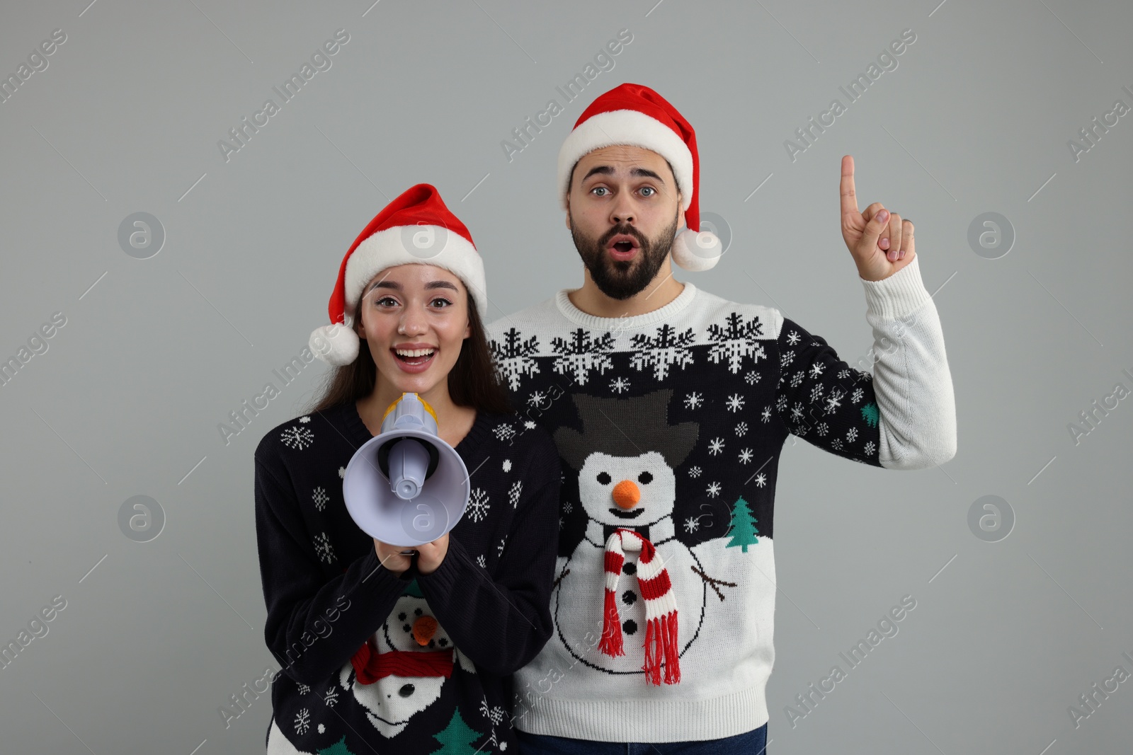 Photo of Young couple in Christmas sweaters and Santa hats shouting in megaphone on grey background