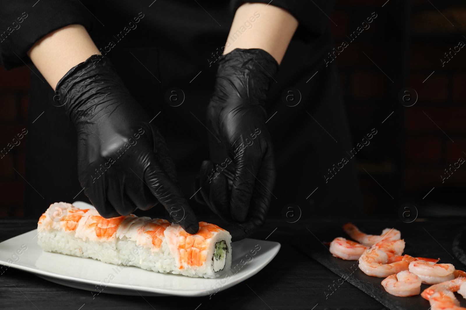 Photo of Chef in gloves making sushi rolls with shrimps at black wooden table, closeup