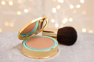 Face bronzer and makeup brush on grey textured table against blurred lights, closeup