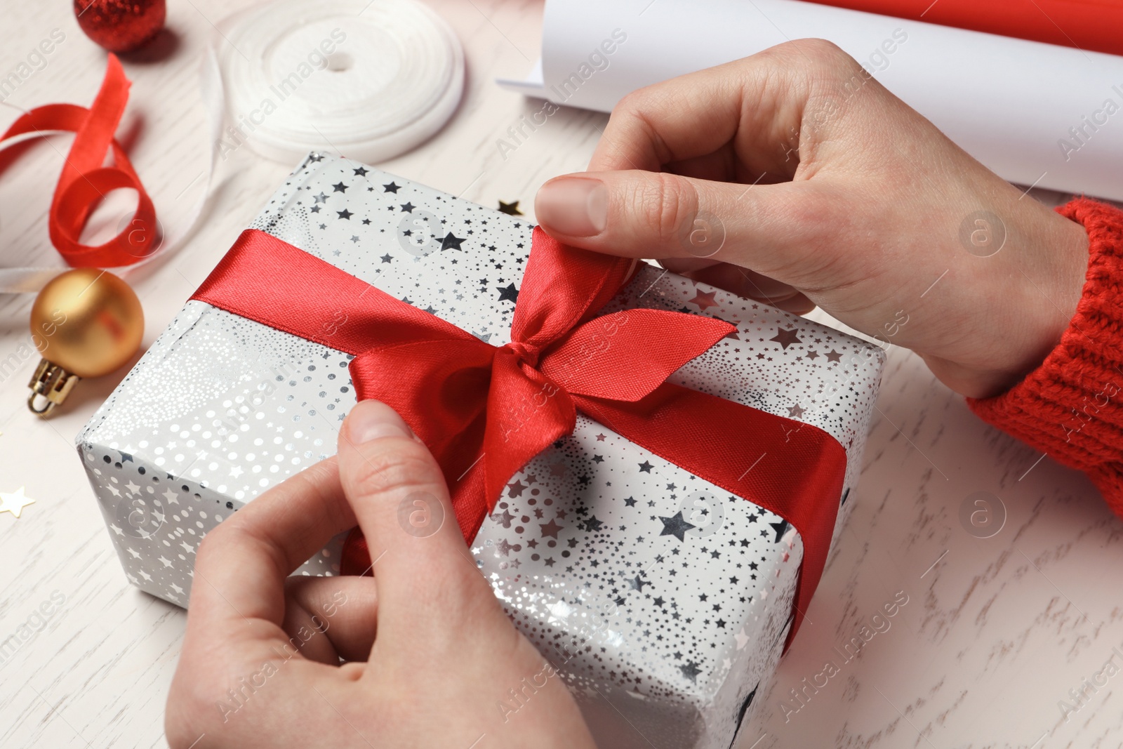 Photo of Christmas present. Woman tying ribbon bow on gift box at white wooden table, closeup