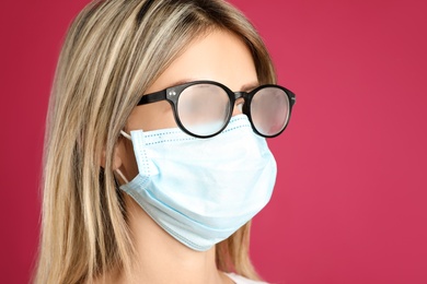 Photo of Woman with foggy glasses caused by wearing disposable mask on pink background. Protective measure during coronavirus pandemic