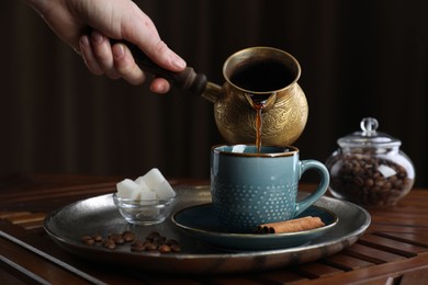 Turkish coffee. Woman pouring brewed beverage from cezve into cup at wooden table, closeup