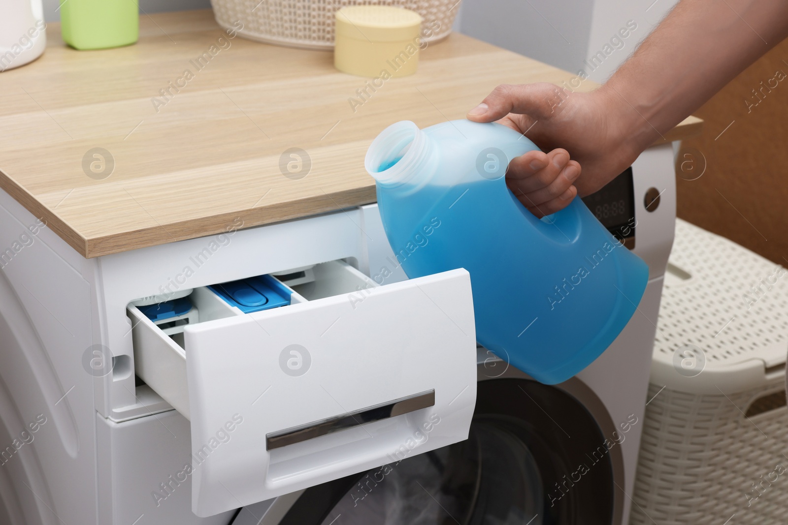 Photo of Man pouring fabric softener from bottle into washing machine indoors, closeup