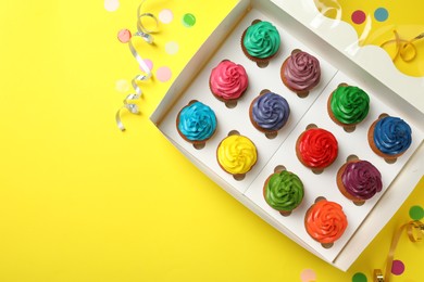 Box with different cupcakes and confetti on yellow background, flat lay. Space for text