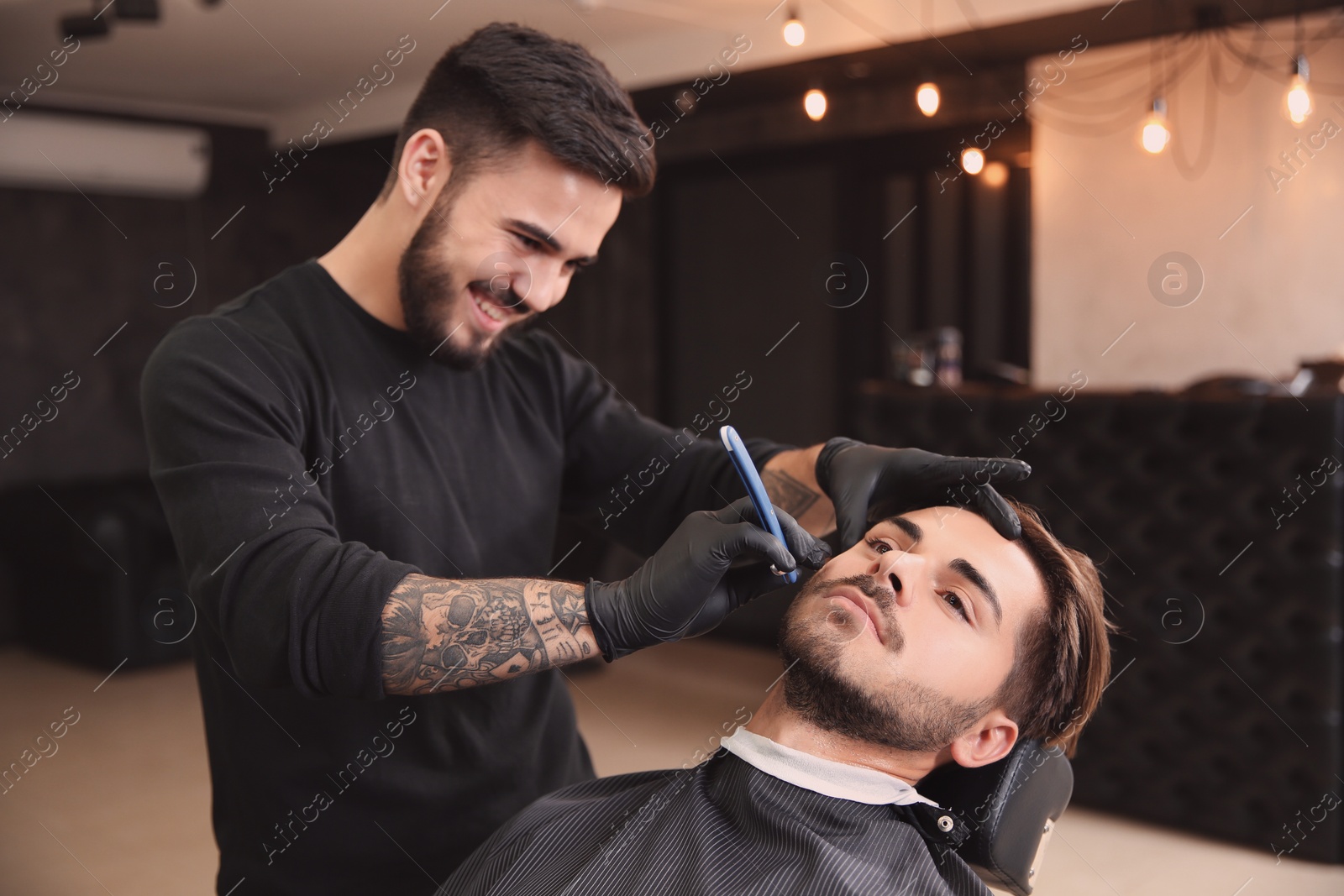 Photo of Professional hairdresser shaving client with straight razor in barbershop