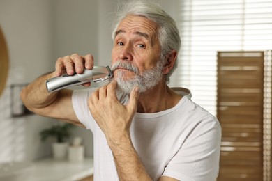 Photo of Senior man trimming beard with electric trimmer in bathroom