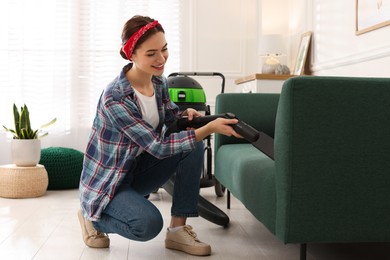Photo of Young woman vacuuming sofa in living room