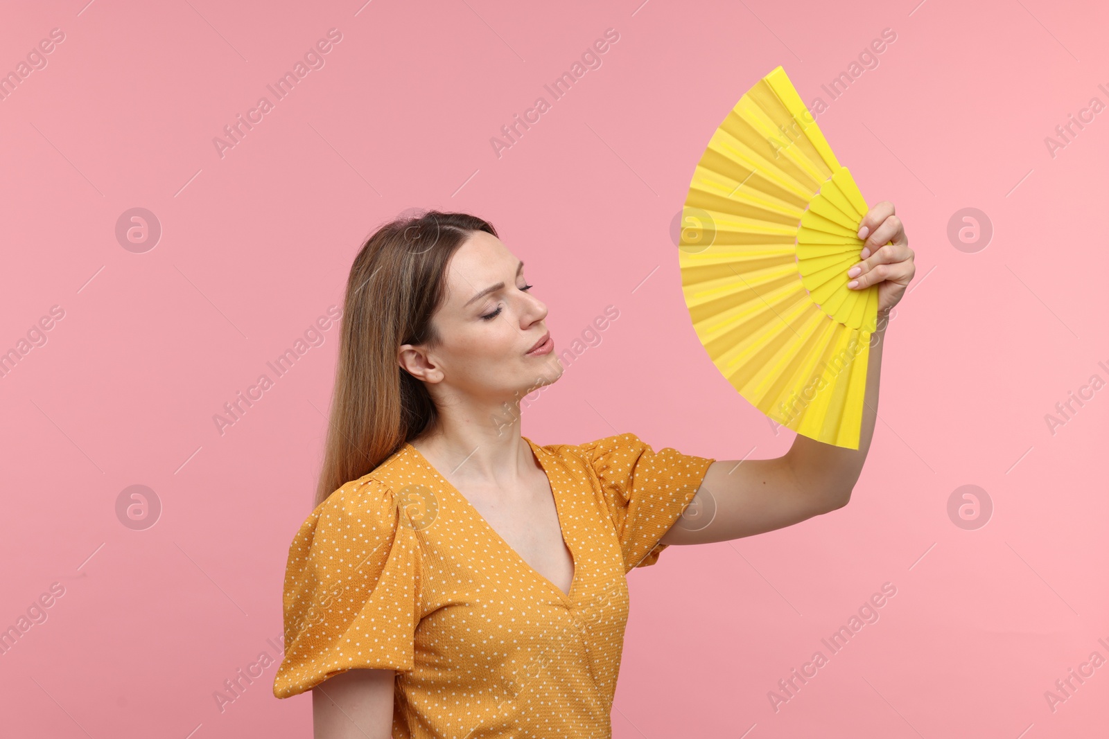 Photo of Beautiful woman waving yellow hand fan to cool herself on pink background