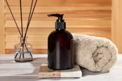 Bottle of shampoo and rolled towel on white table near wooden wall