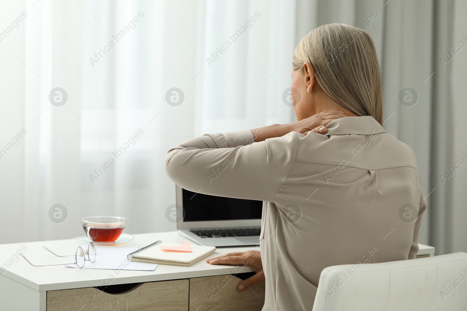 Photo of Woman suffering from neck pain at workplace in room