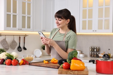 Happy young housewife using smartphone while cooking at white marble table in kitchen