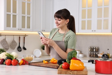 Photo of Happy young housewife using smartphone while cooking at white marble table in kitchen