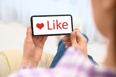 Image of Woman holding smartphone with word Like on screen indoors, closeup