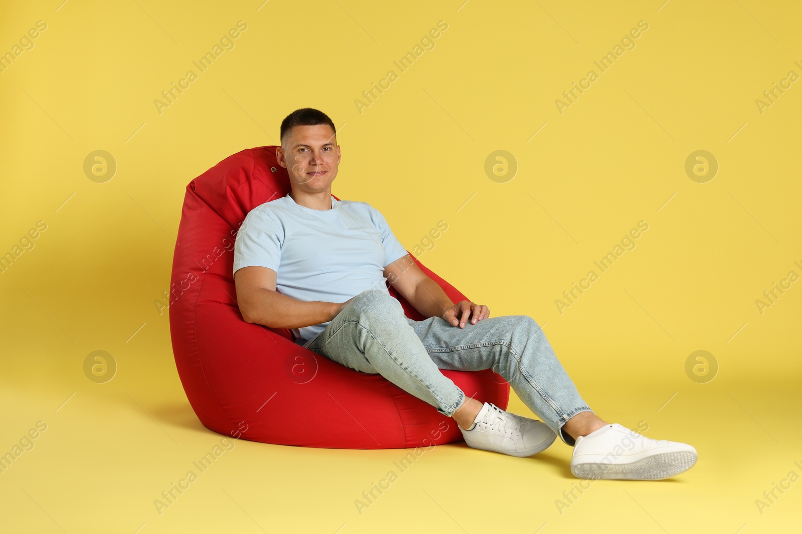 Photo of Handsome man resting on red bean bag chair against yellow background