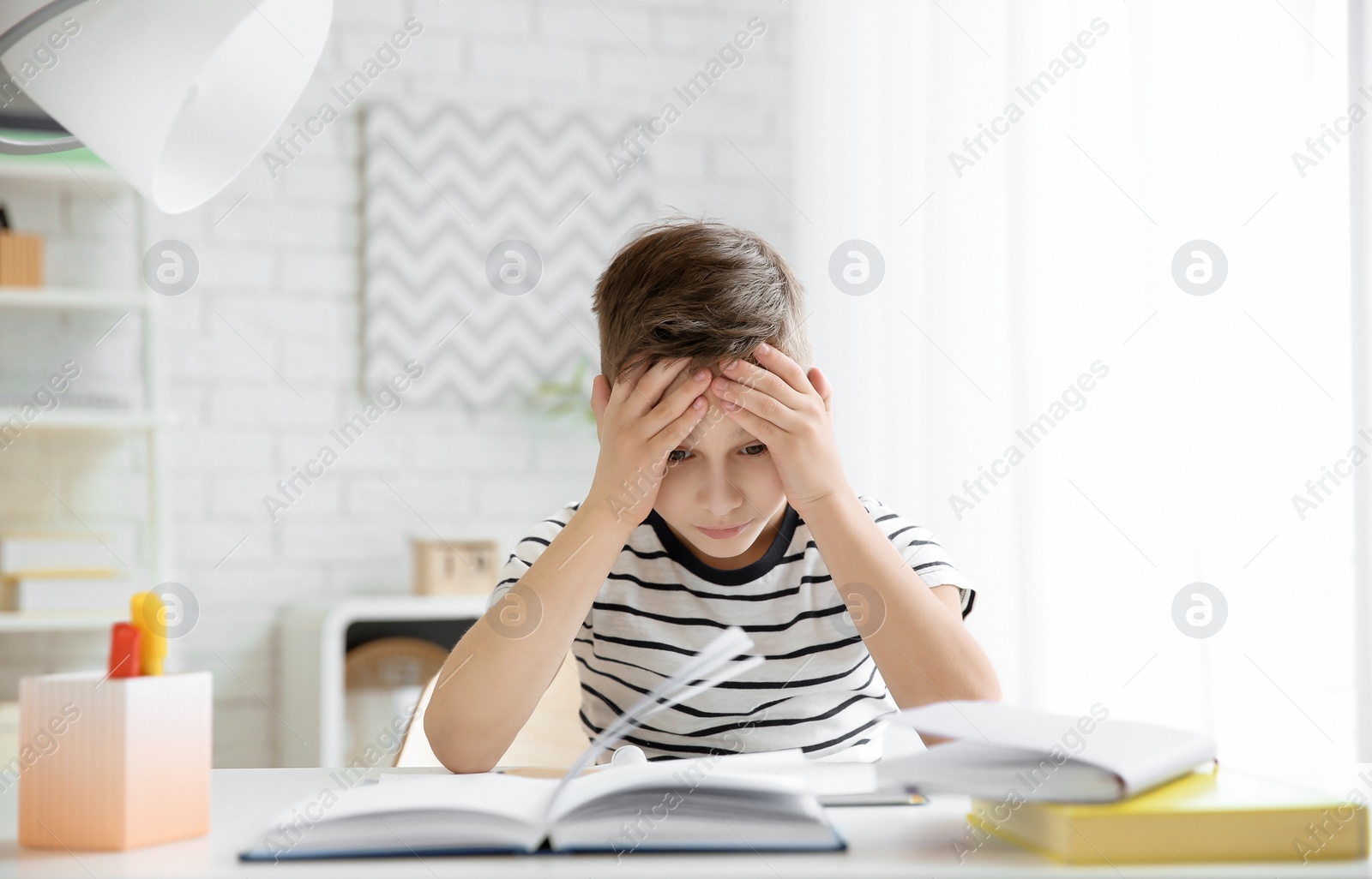 Photo of Little boy suffering from headache while doing homework at table indoors