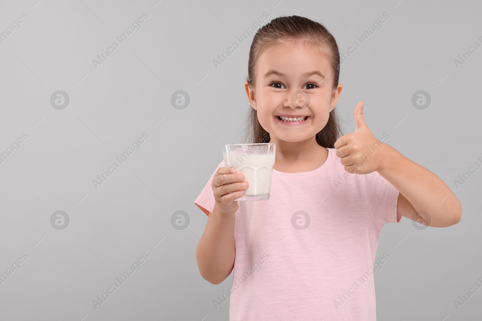 Photo of Cute girl with glass of fresh milk showing thumb up on light grey background, space for text