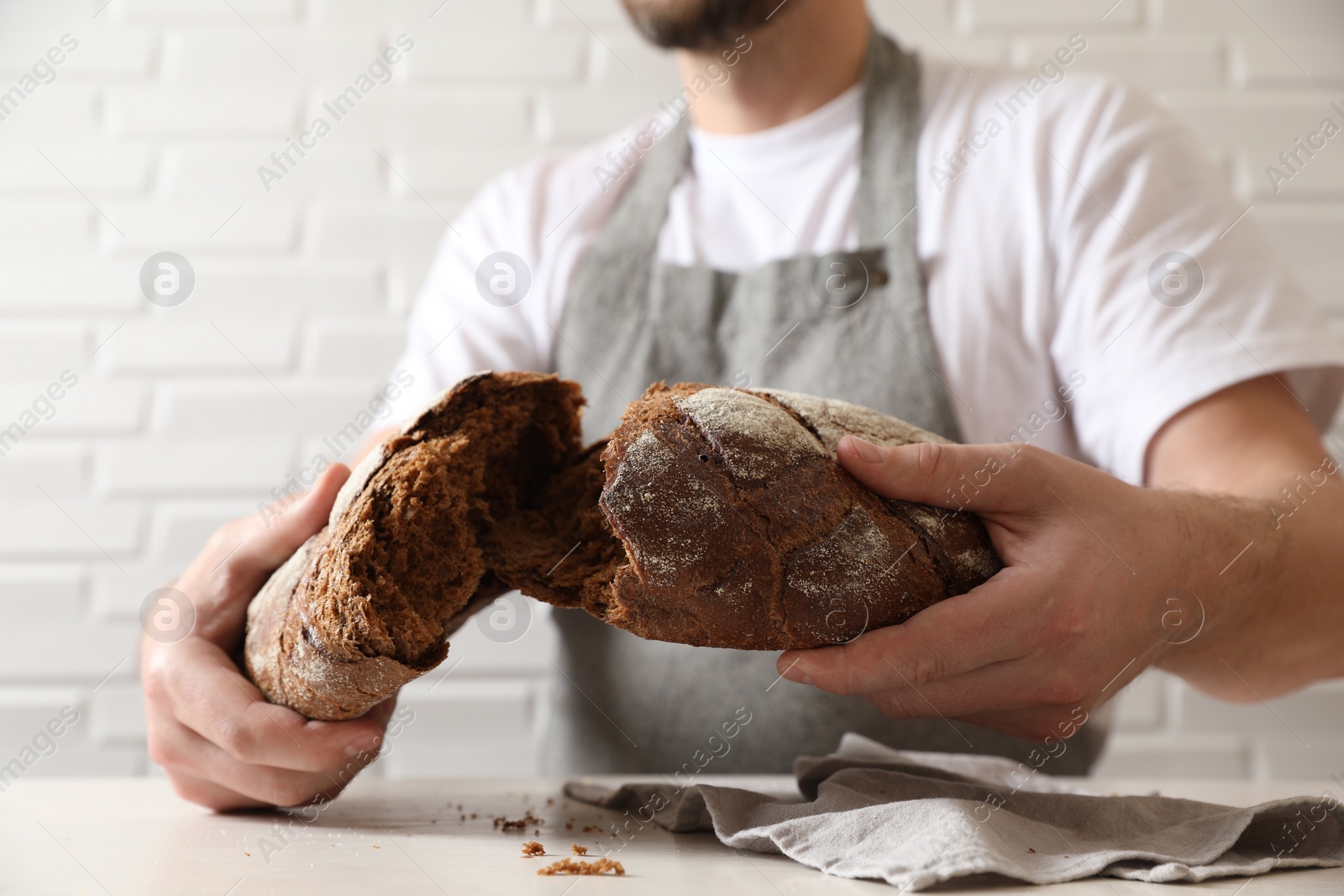 Photo of Man breaking loaf of fresh bread at white table near brick wall, closeup