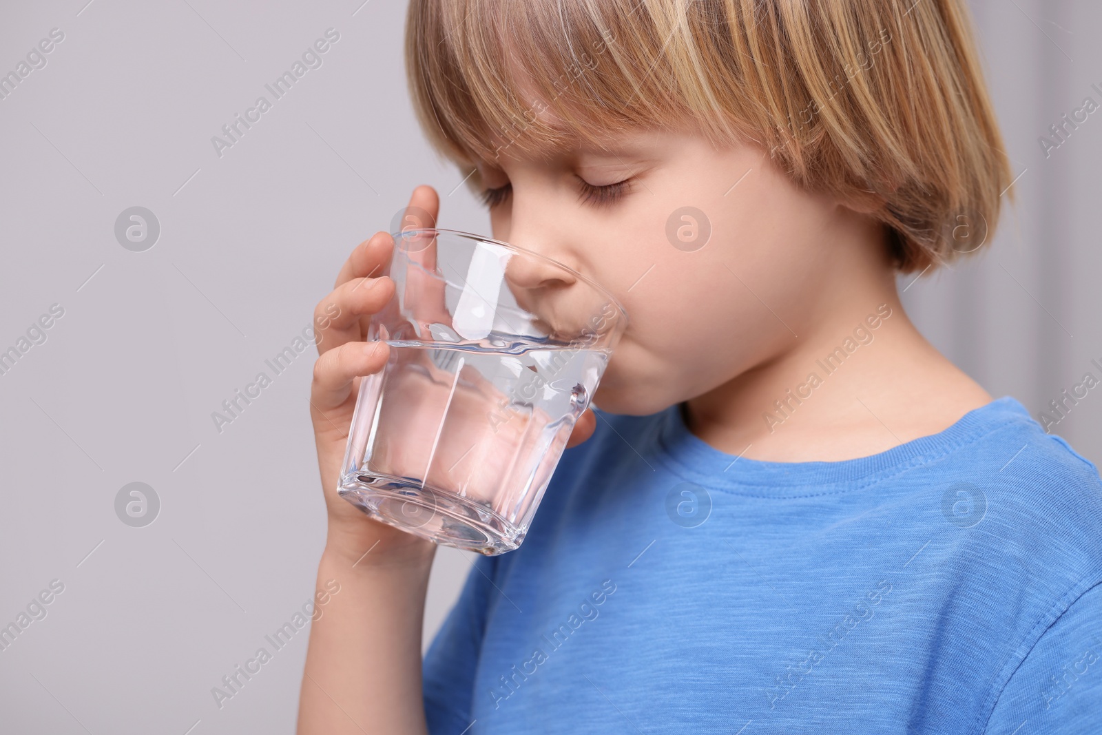 Photo of Cute little boy drinking fresh water from glass on grey background, closeup