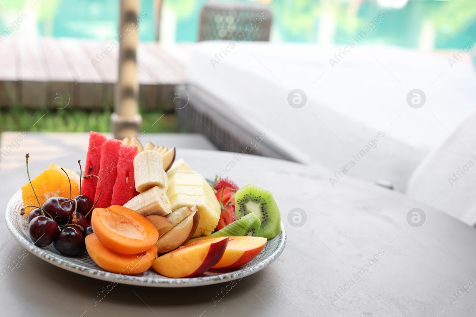 Photo of Plate with fresh fruits on table near sun lounger, space for text. Luxury resort with outdoor swimming pool