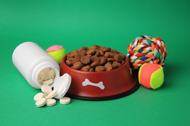 Photo of Bowl with dry pet food, bottle of vitamins and toys on green background