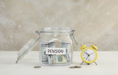 Photo of Glass jar with word Pension, dollar banknotes, coins and alarm clock on white table