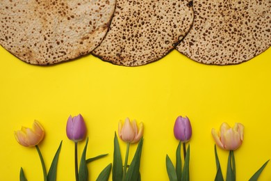 Photo of Tasty matzos and fresh flowers on yellow background, flat lay. Passover (Pesach) celebration