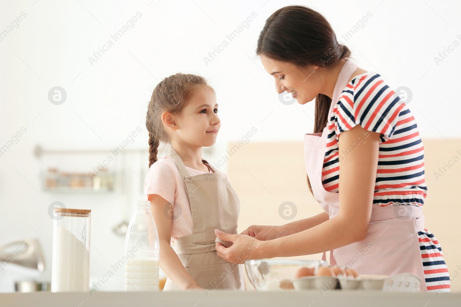 Photo of Young woman helping her daughter to put on apron in kitchen. Making dough together