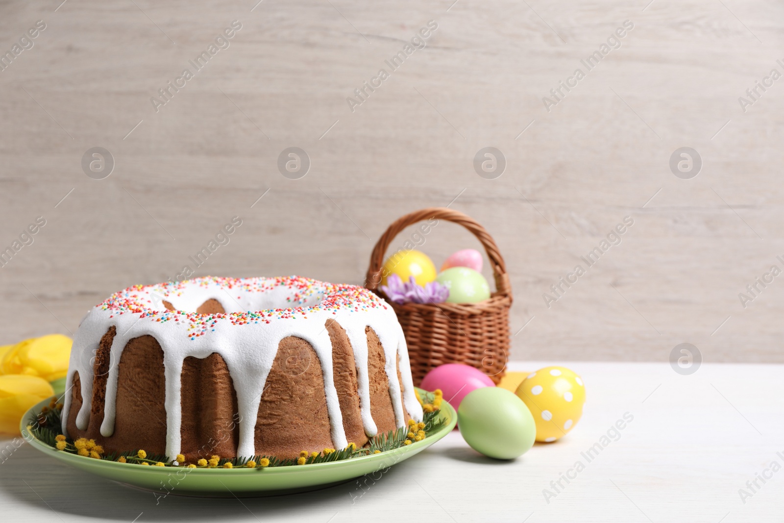 Photo of Glazed Easter cake with sprinkles, painted eggs and flowers on white wooden table, space for text