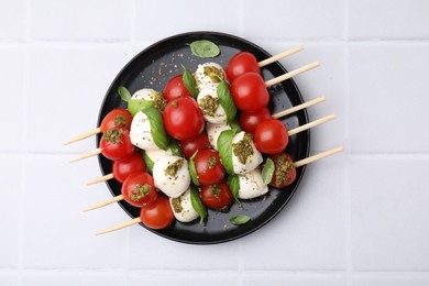 Photo of Caprese skewers with tomatoes, mozzarella balls, basil and pesto sauce on white tiled table, top view
