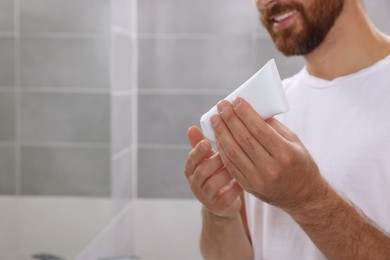 Photo of Handsome man with tube of cream in bathroom, closeup. Space for text
