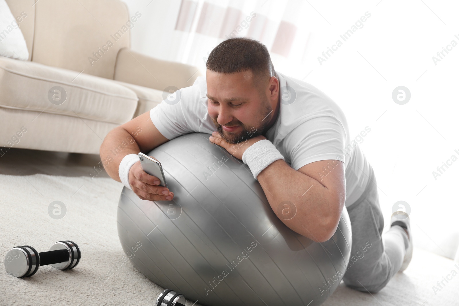 Photo of Lazy overweight man using smartphone while lying on exercise ball at home