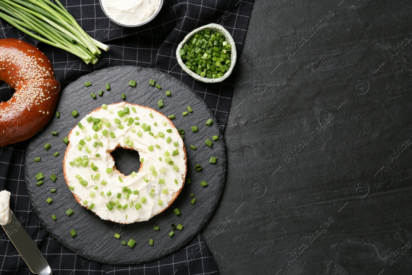 Photo of Delicious bagel with cream cheese and green onion on black table, flat lay. Space for text