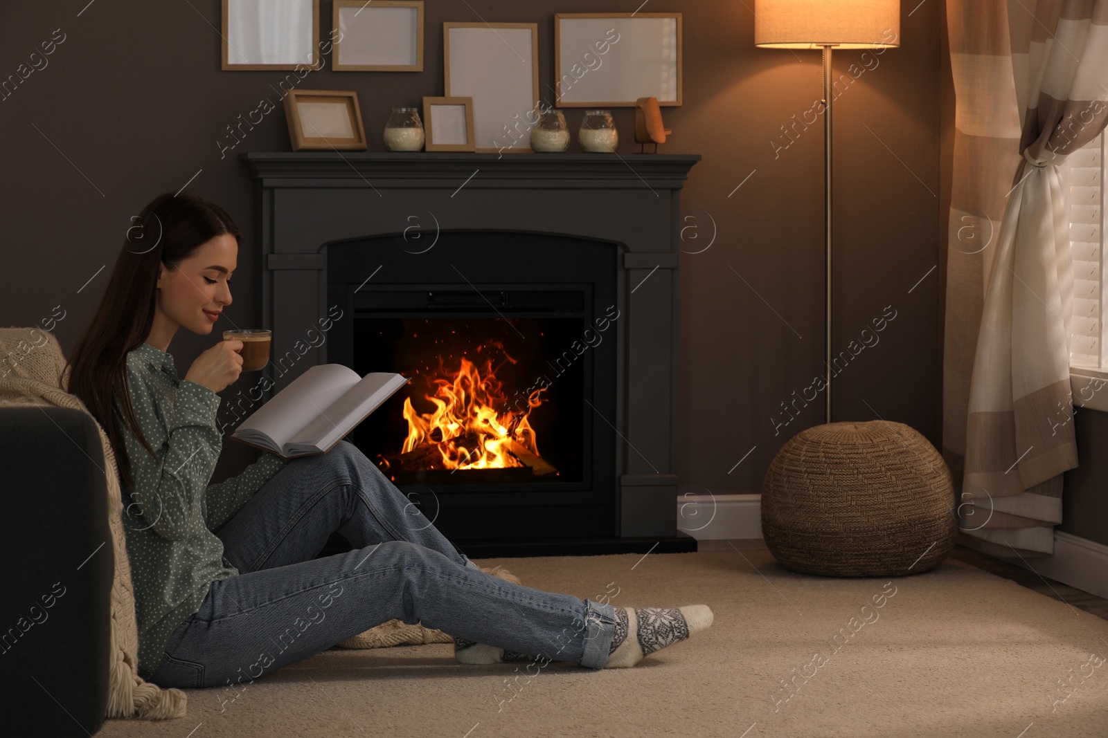 Photo of Beautiful young woman with cup of coffee reading book on floor near fireplace at home. Space for text