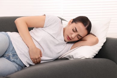 Photo of Woman suffering from abdominal pain while lying on sofa indoors. Unhealthy stomach