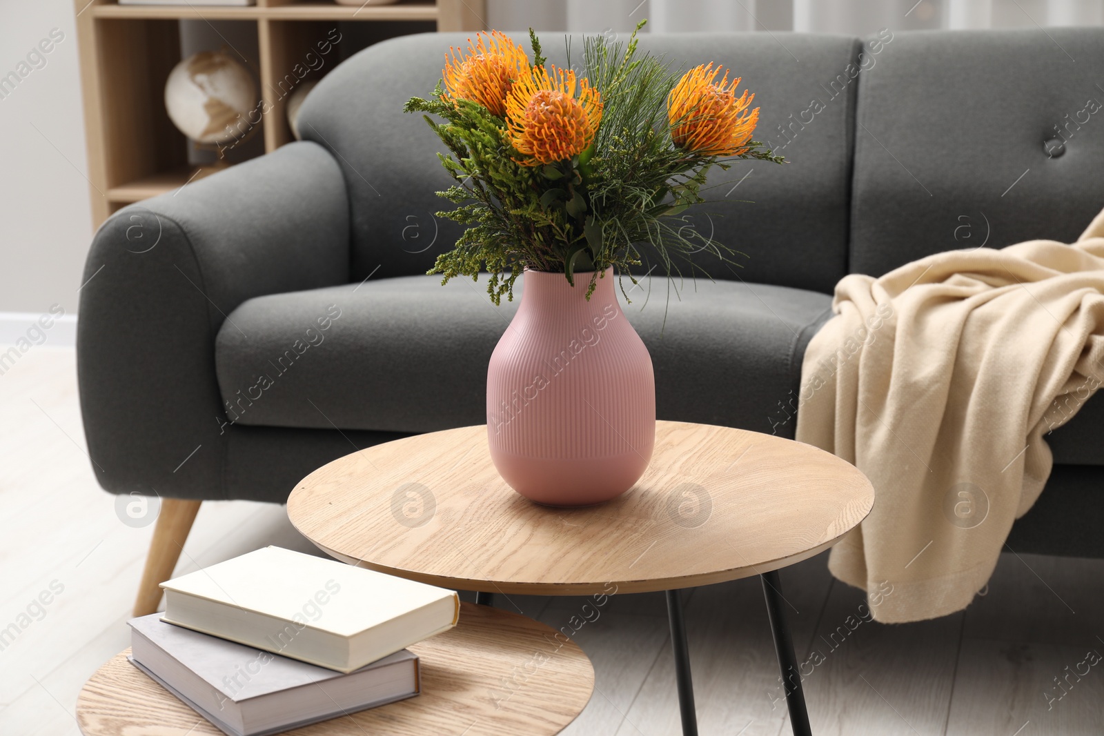 Photo of Vase with beautiful flowers and books on wooden nesting tables near grey sofa indoors