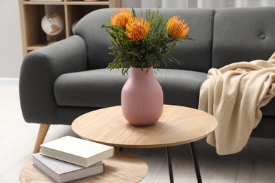 Vase with beautiful flowers and books on wooden nesting tables near grey sofa indoors