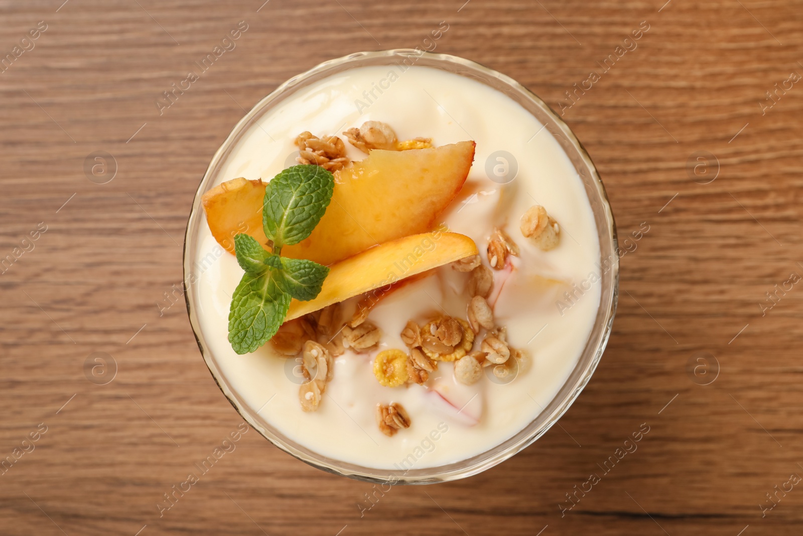 Photo of Tasty peach yogurt with granola, mint and pieces of fruit in dessert bowl on wooden table, top view